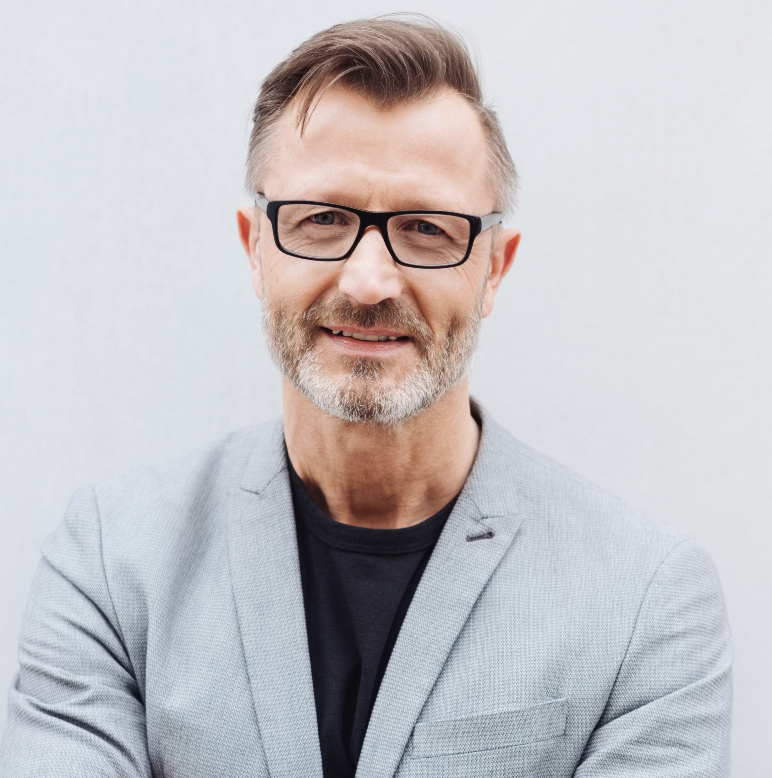 Portrait of smiling mature bearded man wearing glasses standing with arms crossed against bright background